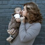 Szkoła Językowa Lagarta Loving mother kisses her baby, showcasing a tender family moment against a dark brick wall.