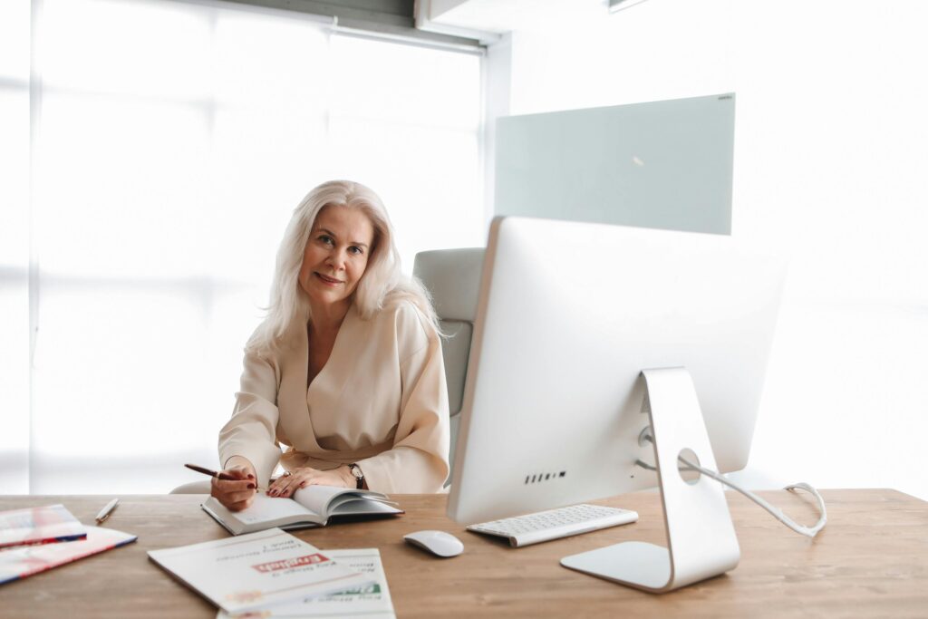 niemiecki online Elegant senior woman writing at office desk with computer, showcasing a professional work environment.